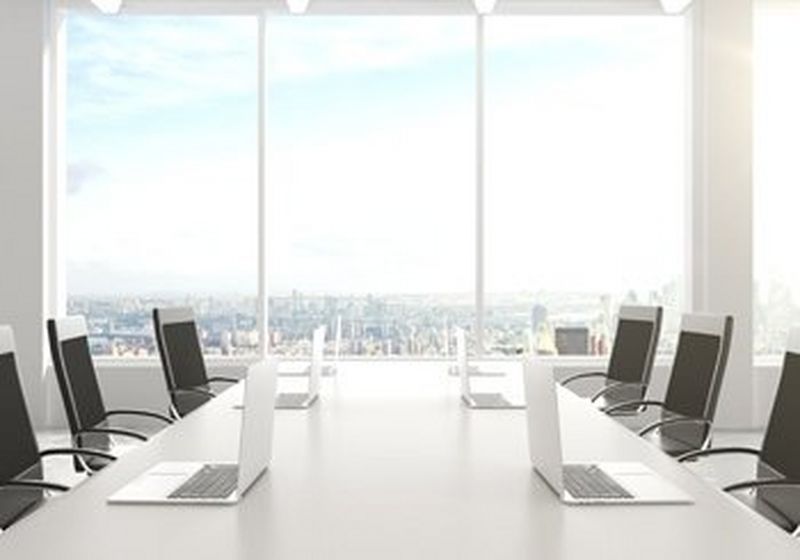 A boardroom table with six chairs and six laptops, against a backdrop of a window, overlooking a city. 