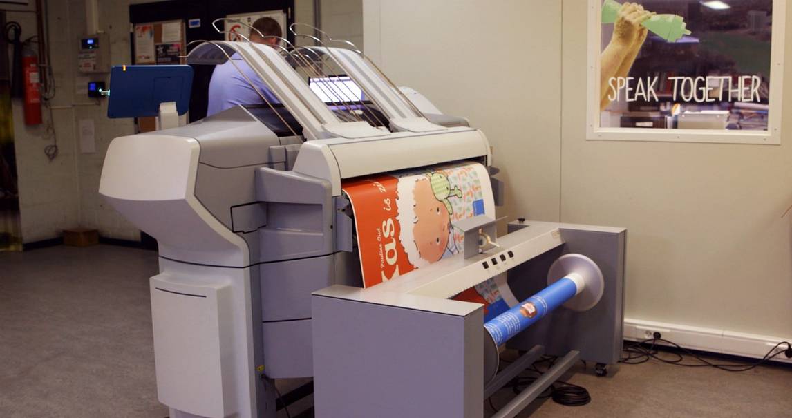 Commercial digital printer in an office prints an A1 sheet of paper with a cartoon image of a child on an orange background.