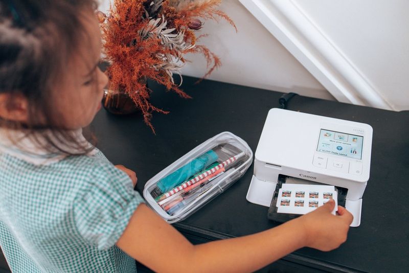 A young girl prints a sheet of mini stickers from a Canon compact printer. Next to the printer is a pencil case filled with stationery.