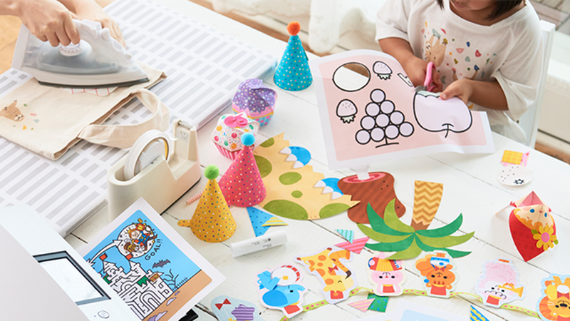 A child cutting out an apple shape from a craft paper sheet, surrounded by different crafts on a table.