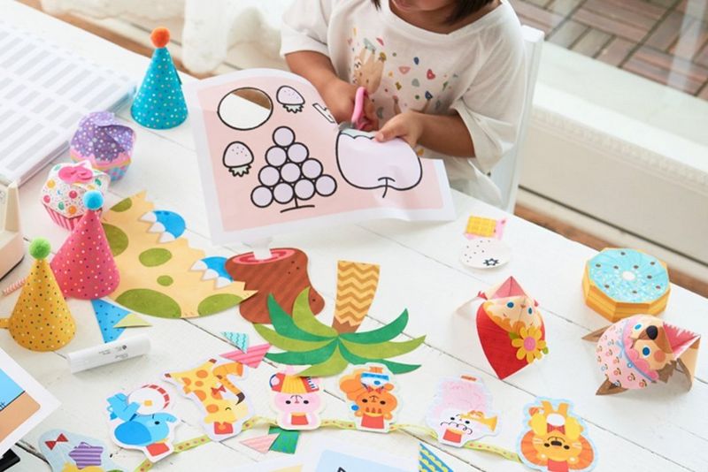 A child cutting out an apple shape from a craft paper sheet, surrounded by different crafts on a table.