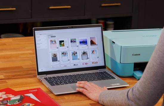 A laptop sits open on a desk with the Canon Easy-PhotoPrint software onscreen, Canon paper to the left and a Canon printer to the right.