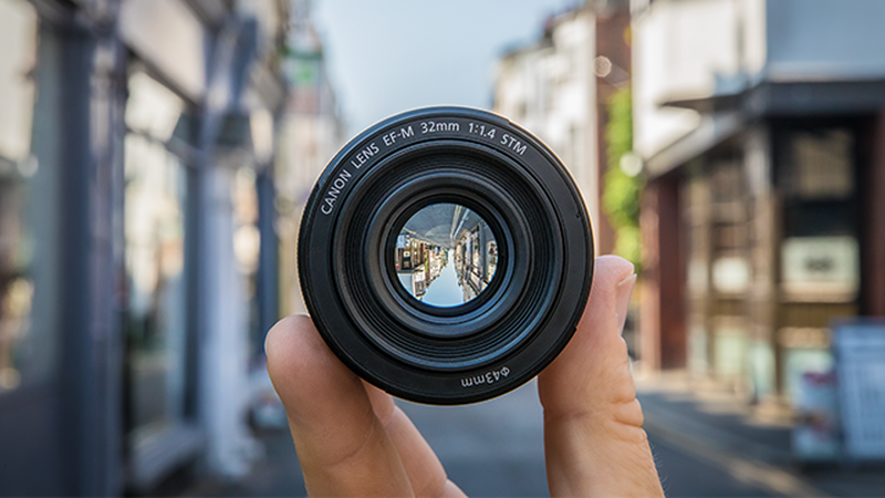 A hand holding lens EF-M 32mm f/1.4 STM in the street showing the same street upside down through the lens.