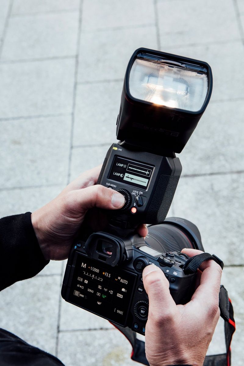 A photographer's hand adjusts the settings on the back of a Canon Speedlite EL-1 attached to an EOS R5 camera held in his other hand.