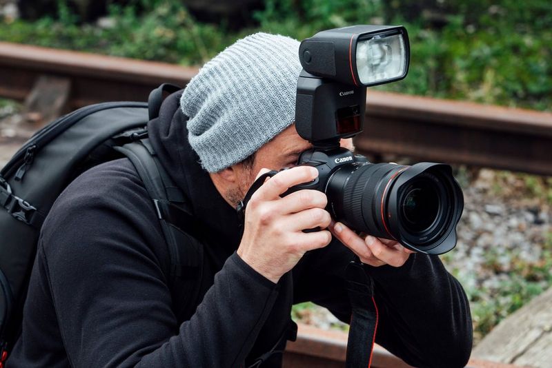 A man crouching down, using a Canon EOS R5 with a Canon Speedlite EL-1 attached in its hot-shoe. 