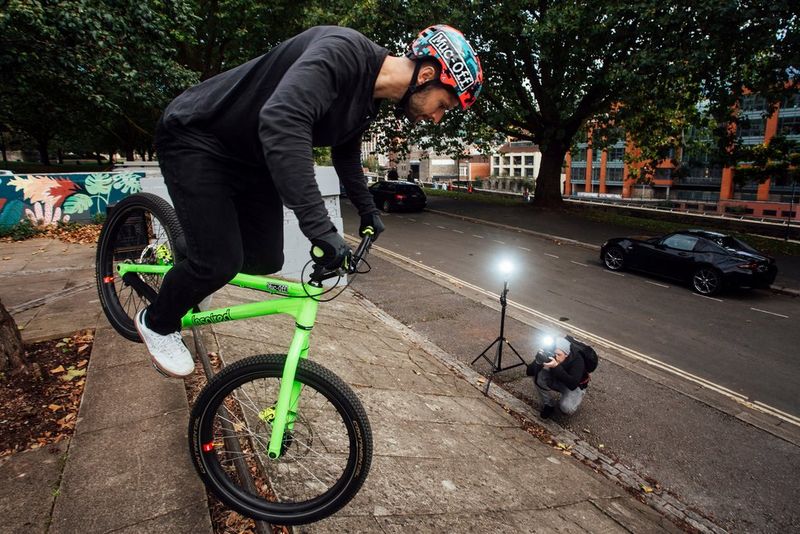 A stunt rider on a bright green bicycle balanced on its front wheel being photographed using two Canon Speedlite EL-1 flashes. 