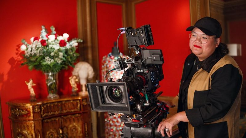 A woman wearing a cap and glasses, dressed in a black and tan jacket, stands behind a Canon EOS C400  setup in a luxurious room.