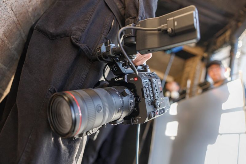 A close-up shot of a professional Canon video camera being held by a person dressed in dark clothing. The camera, equipped with a large lens and a monitor, is focused on capturing a scene. 