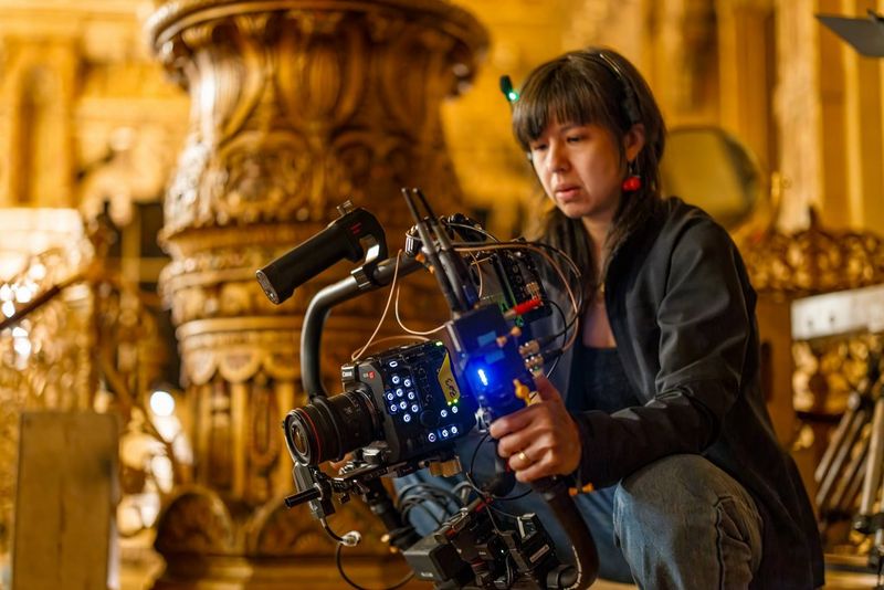 A close-up shot of a handheld D224 camera rig on a film set, with a focus on the detailed controls and wiring of the professional camera. The rig is positioned on a colourful carpet, and the hands of the cinematographer are visible.