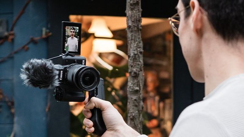 A man holding Canon EOS M50 Mark II with a mic on a tripod in front of him, speaking to the camera.