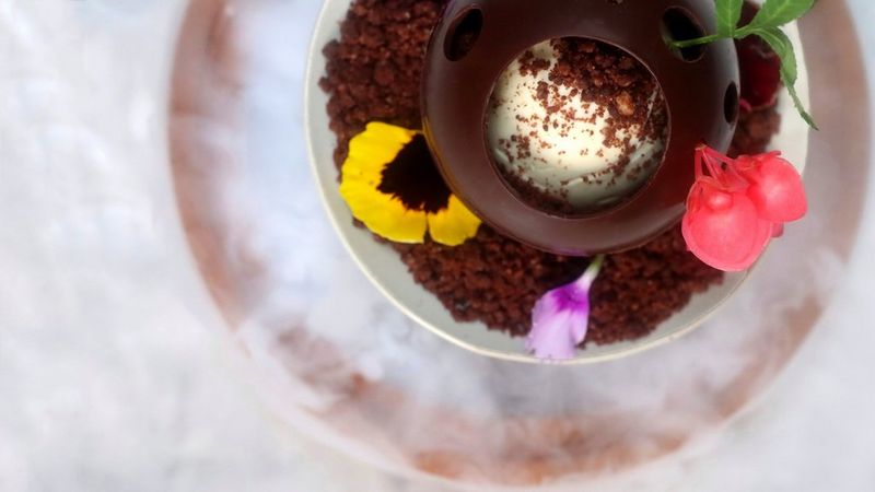 A hollow chocolate ball filled with ice cream and garnished with petals and chocolate sprinkles. 