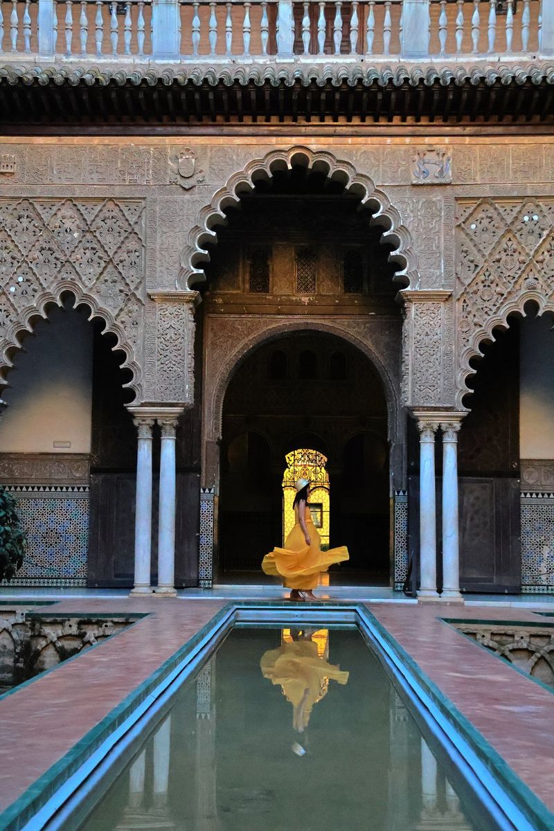 A woman in a yellow dress stands beneath an ornate archway in front of a long, narrow pool of water in which her reflection can be seen.