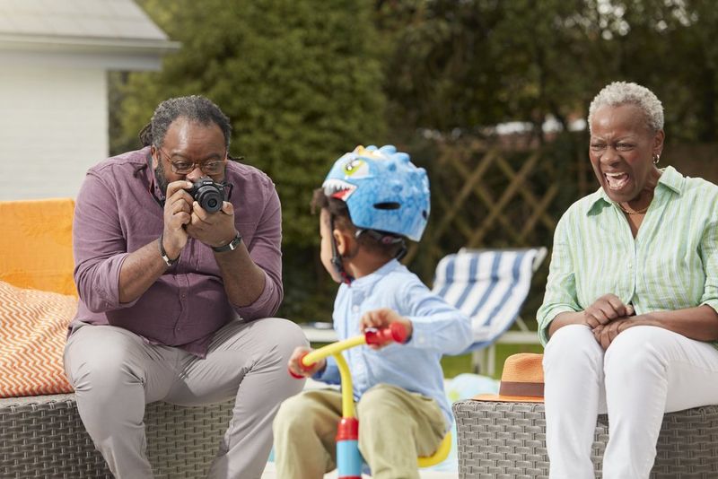 Grandfather taking a picture using Canon EOS R100 of his grandchild riding a bike. Grandmother is sitting close by smiling.