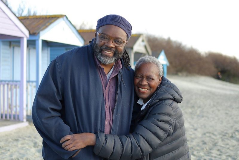 Two people stand with their arms around each other, smiling, in front of a row of beach huts on the sand, taken on a Canon EOS R100.