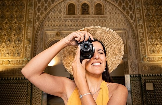 A woman in a summer dress and sunhat holds the Canon EOS R10 up to her face to look through the viewfinder, as she stands in front of detailed Spanish architecture.