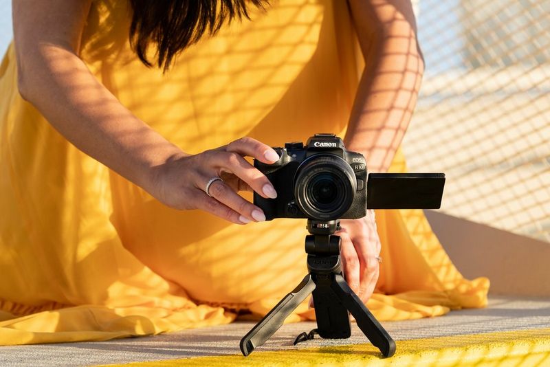 A woman in a flowing yellow dress crouches down to adjust the position of a Canon EOS R10 on a small tripod.