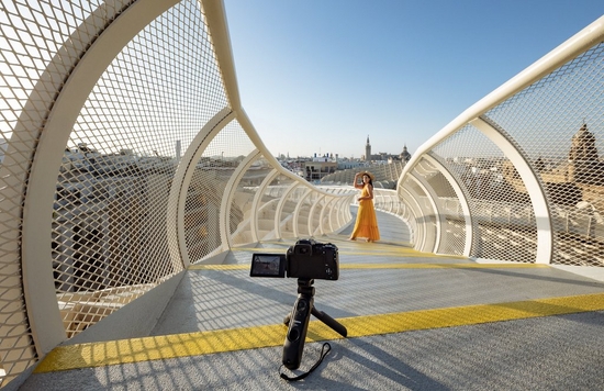 A woman in a yellow sundress poses on a curving footbridge, with a Canon EOS R10 on a small tripod set up to photograph her.