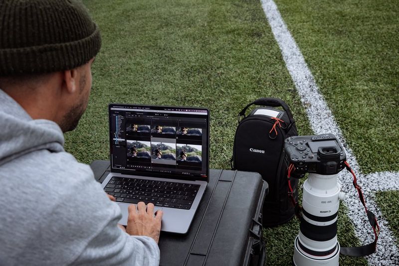 A photographer works on a laptop on a football field with the Canon EOS R1 camera beside them, lens down on the grass.
