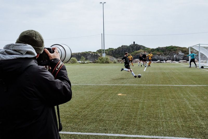A photographer stands on the sidelines holding the Canon EOS R1 camera capturing a game of football in action. 