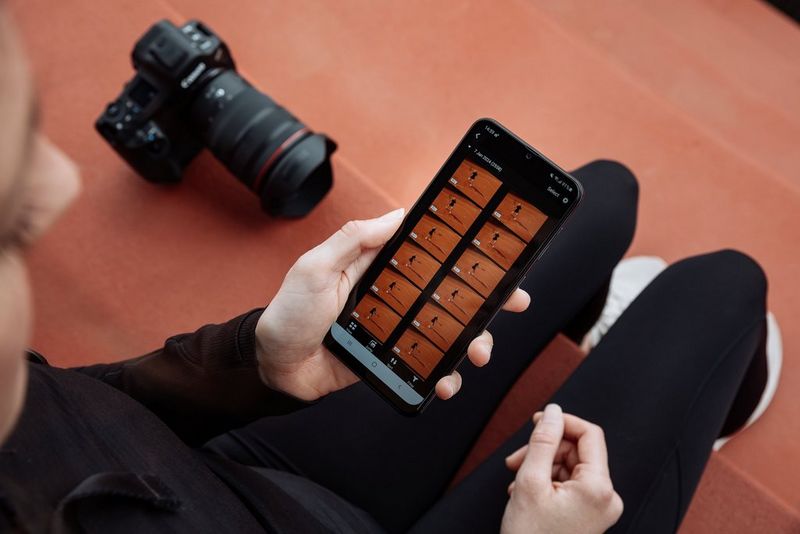 A hand holding a smartphone running the Canon Camera Connect app, with the Canon EOS R1 camera sitting alongside on an orange tarmac step.