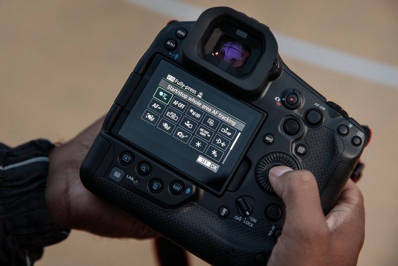 A photographer's hand holds a Canon EOS R1 camera with the AF-ON button customisation screen.  