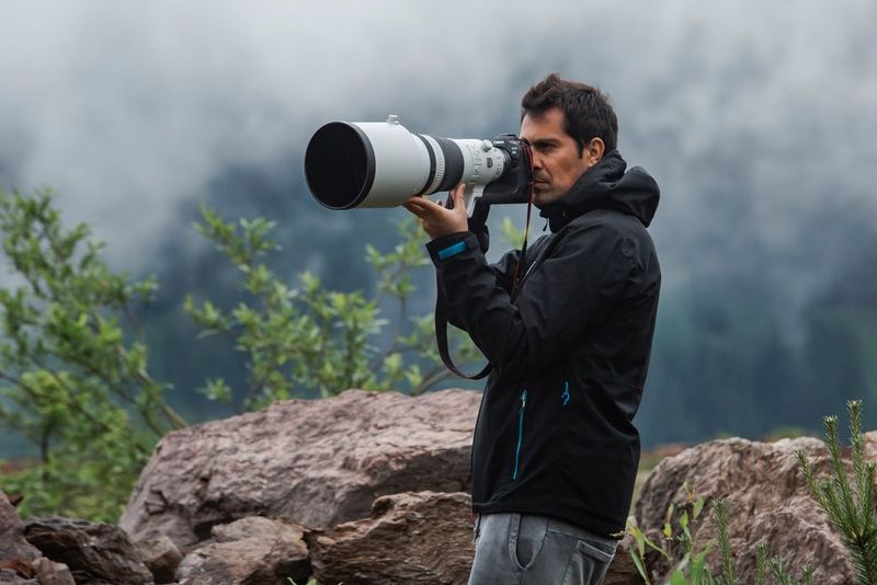 A man in a black jacket shoots handheld with a Canon EOS R3 and a super-telephoto lens, low clouds visible above the rocks behind him.