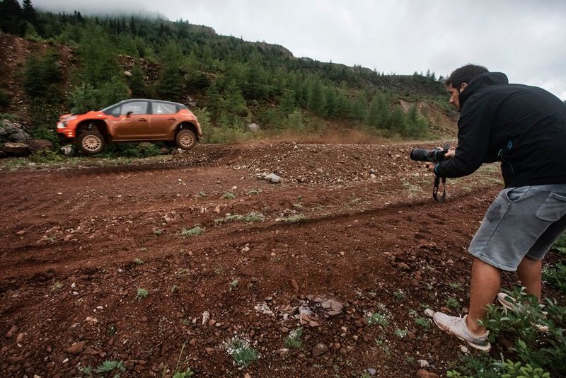 A man wearing shorts and a dark jacket photographs an electric rally car speeding through rocky terrain. 