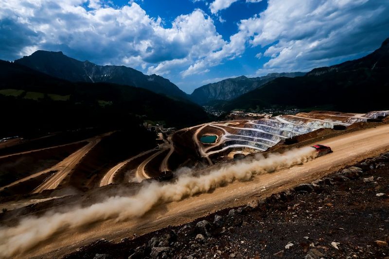 A red rally car kicks up a long trail of dust as it travels at speed along a dirt road above a quarry.