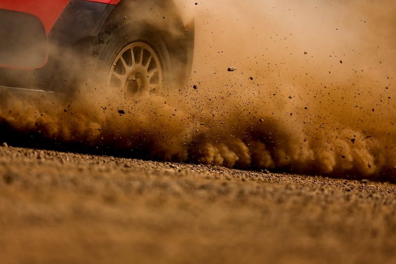 A close-up of the rear wheel of a red rally car as it kicks up plumes of dust and dirt from the road.