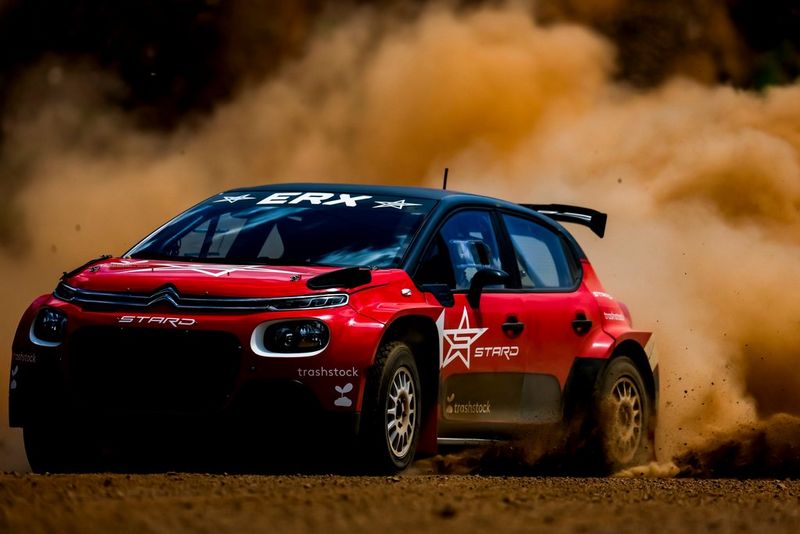A close-up of a red and black sports car on a dusty road, a cloud of dust and dirt spraying up behind it.