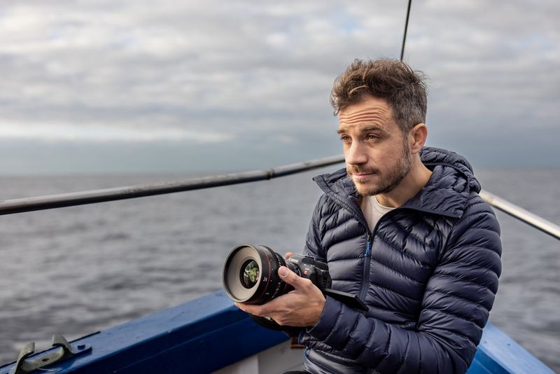 A man in a blue jacket filming with an EOS R5 C camera on a boat in choppy waters. 