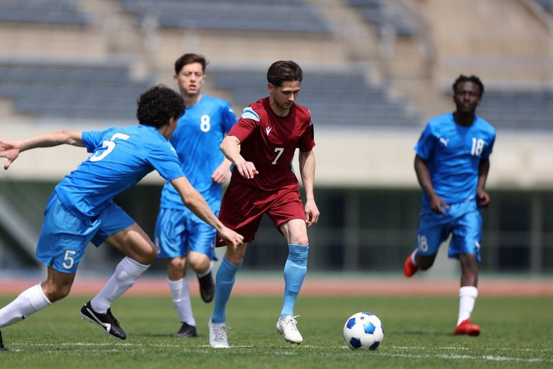  Four men play football in a stadium, with three players in blue kit surrounding one man in a red kit in possession of the ball. Shot on Canon EOS R5 Mark II. 