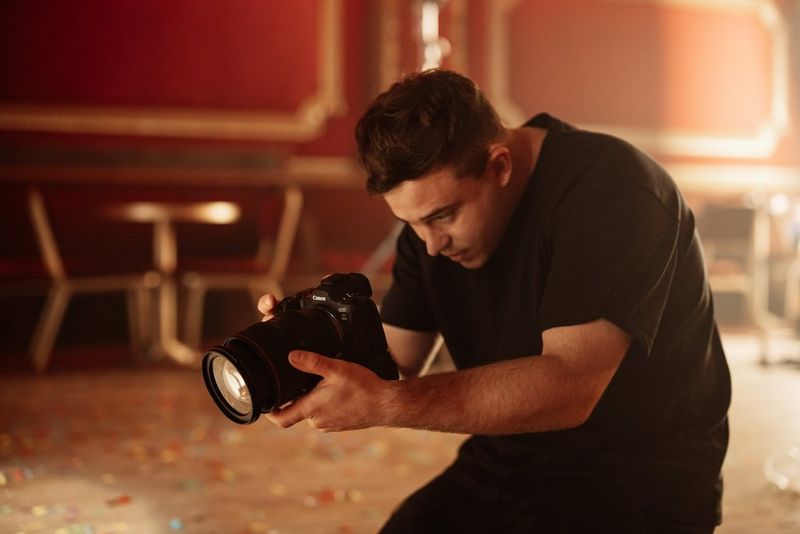 A photographer stands in a hunched position holding the Canon EOS R5 Mark II camera with a blurry background of music revenue in the background. 