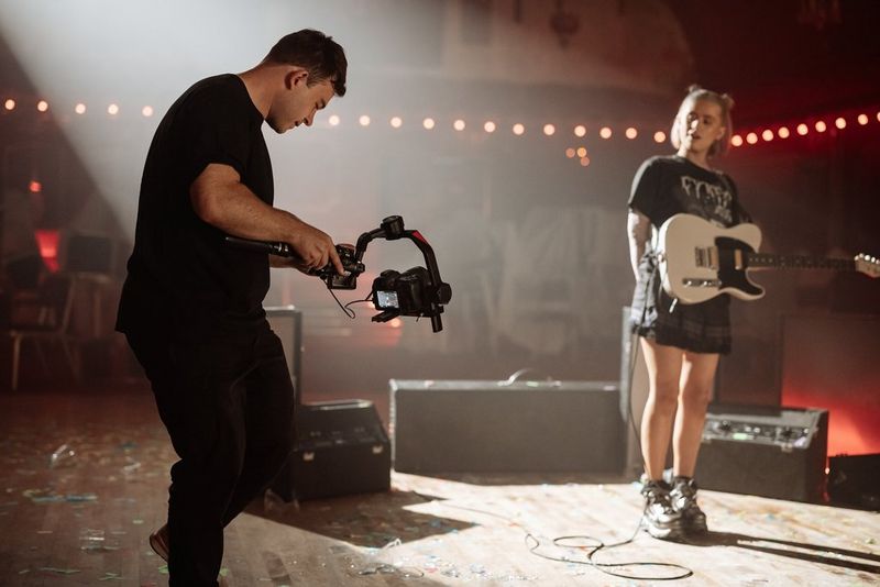 A photographer holding a gimbal with the Canon Canon EOS R5 Mark II attached shooting a girl with a guitar strapped around her body on stage.