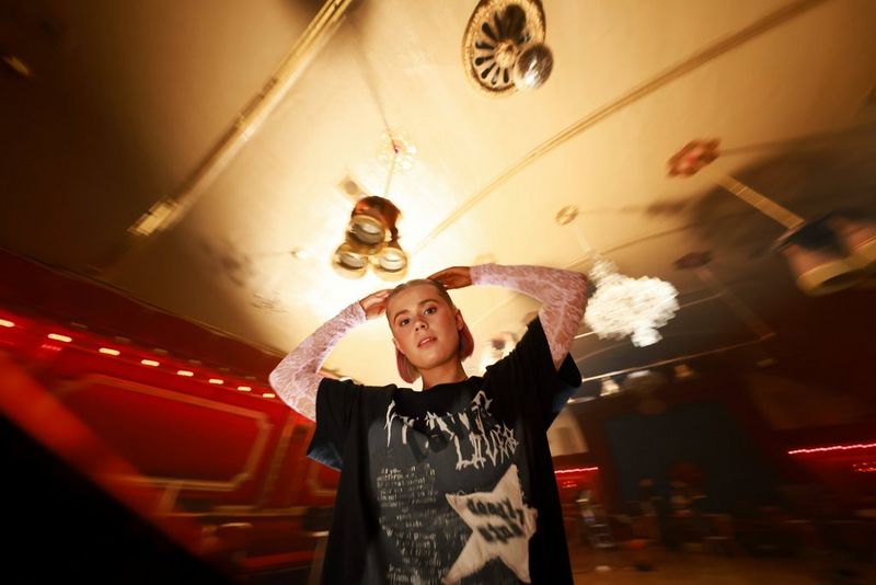 A girl stands in a music venue looking down the lens with her hands on her head with a slightly distorted and blurry background. Shot on Canon EOS R5 Mark II.  