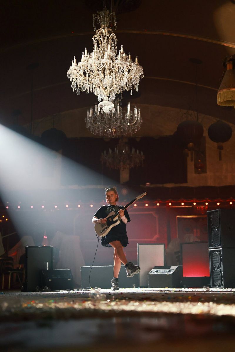 A girl standing on a stage playing a guitar with a chandelier positioned above her head with an unknown spotlight coming into the frame from the left. Shot on Canon EOS R5 Mark II.