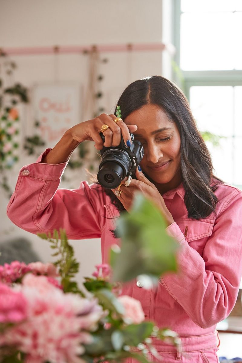 A woman in a pink jumpsuit filming a floral display with a Canon EOS R50 camera. 