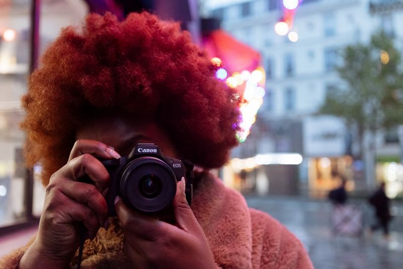 Content creator Fatou N'Diaye stands in a dimly lit street holding a Canon EOS R50 up to her face and looking into the viewfinder.