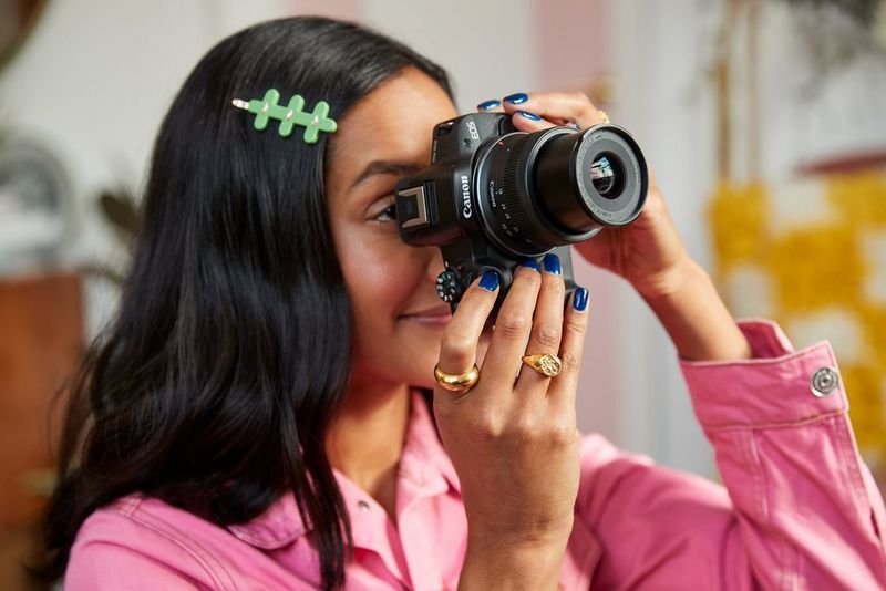 A woman holding a Canon EOS R50 towards their face and looking through the electronic viewfinder.