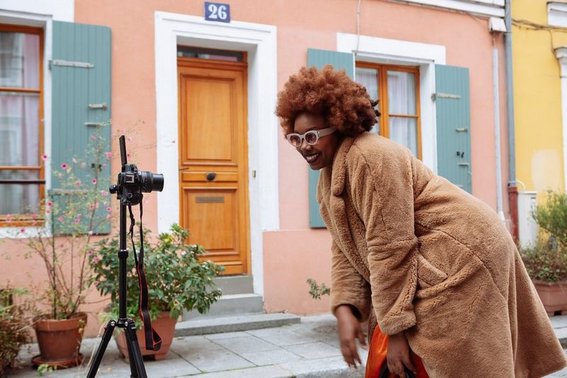 Socialmedia-influencer Fatou N'Diaye, in een pluizige bruine jas, zit gehurkt voor een Canon EOS R50-camera op een statief en glimlacht.