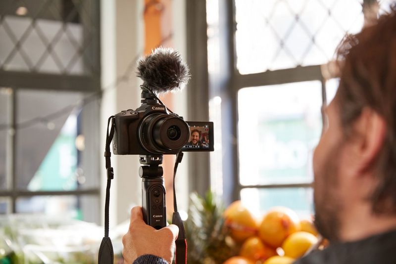 A hand holding a Canon Tripod Grip HG-100TBR attached to a Canon EOS R50 with a Canon Stereo Microphone DM-E100.