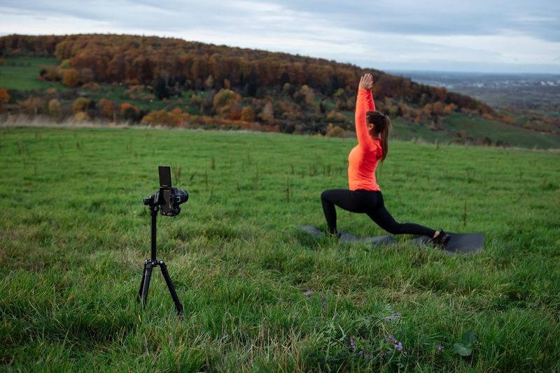 A Canon EOS R50 on a tripod filming content creator Elena Bulkowski in an orange top performing a yoga pose in a field.
