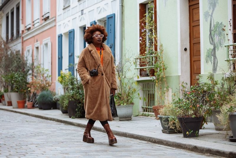Social media influencer Fatou N'Diaye, wearing a fluffy brown coat and glasses and carrying a Canon EOS R50, walks past pastel-coloured houses.
