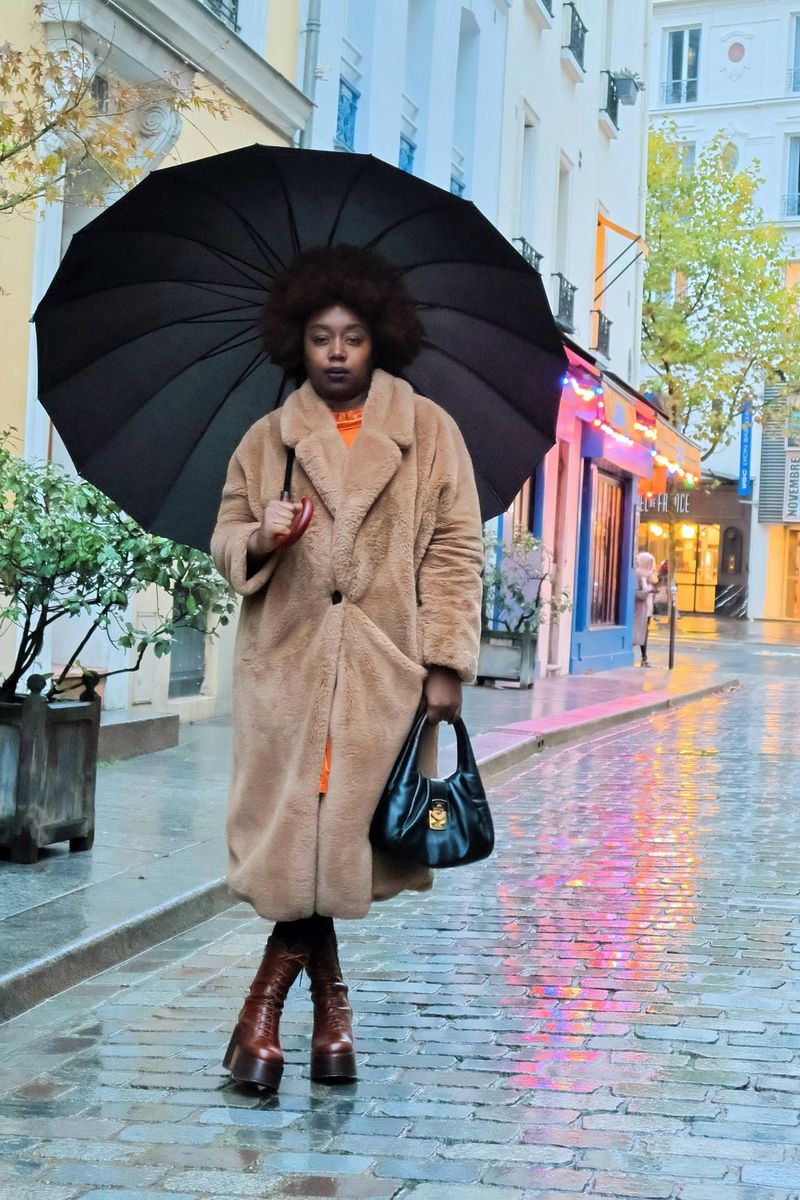 Social media influencer Fatou N'Diaye, wearing a fluffy brown coat and holding an umbrella, stands in a road next to illuminated shopfronts.