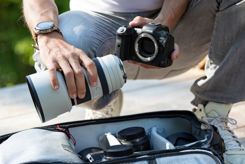 A man holds a Canon EOS R5 in one hand and a Canon telephoto lens in the other, above a kitbag containing other lenses.