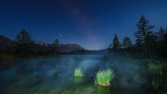 Ein schimmernder See unter einem Sternenhimmel, bei wenig Licht fotografiert. Im Vordergrund sind zwei Schilfgürtel und im Hintergrund ein weitläufiges Gebirge zu sehen.