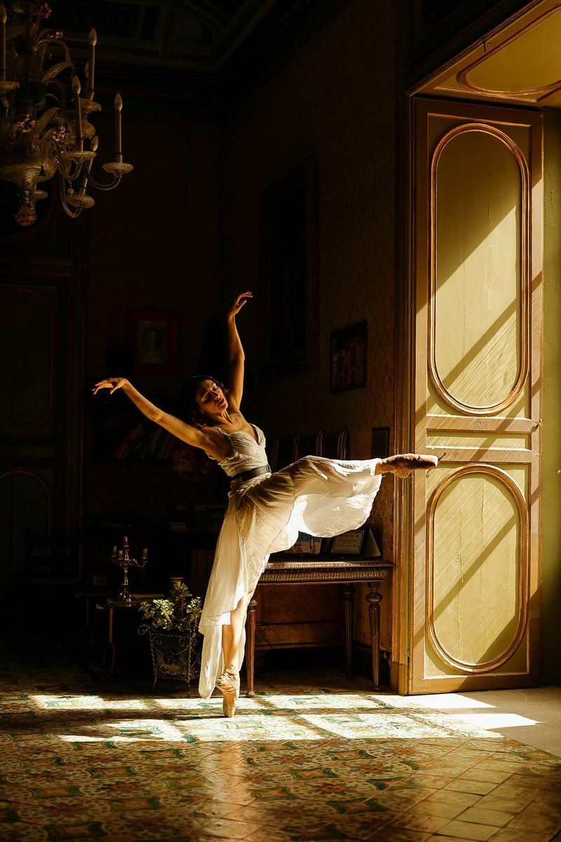 A ballet dancer balances on one leg with her arms outstretched as sunlight streams through the shutters in front of her. Taken on a Canon EOS R6 with a Canon RF 50mm F1.2L USM lens. 