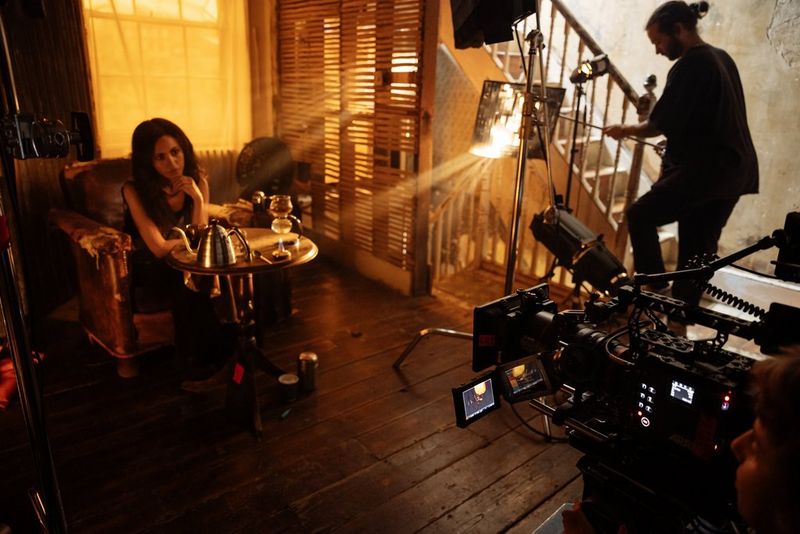 A person sits in an armchair, light coming through shutters and the scene bathed in an ochre glow, a Canon camera and lens pointed at the scene from the right.