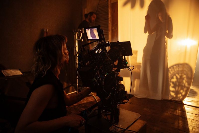 Diana Olifirova sits behind a Canon camera filming a figure in silhouette behind a curtain. 
