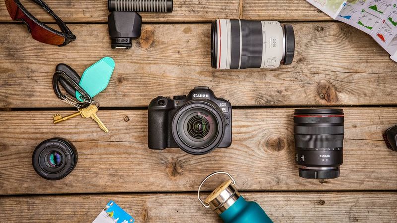 A top-down shot of a Canon EOS R6 Mark II on a wooden surface, surrounded by Canon lenses, sunglasses, a map, a set of keys and a water bottle.